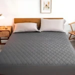 Protector de colchón antifluido color gris con textura acolchada en rombos, exhibido en cama con cabecero de madera y almohadas blancas.