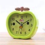 Reloj despertador verde en forma de manzana con manecillas negras y dos caritas sonrientes.
