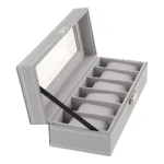 Relojero X6 en color gris claro, con cristal transparente en la tapa y seis compartimentos internos para almacenamiento de relojes. Incluye llave de seguridad visible en el lateral derecho.