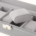 Relojero X6 en color gris claro, con compartimentos internos acolchados y un reposabrazos central para exhibición de relojes. Incluye cierre metálico con llave visible en la parte frontal.