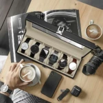 Relojero X6 negro con tapa de vidrio, abierto sobre mesa de madera, mostrando seis compartimentos con relojes de diferentes estilos y colores. Incluye teléfono móvil, cámara, taza de café y revista al fondo.