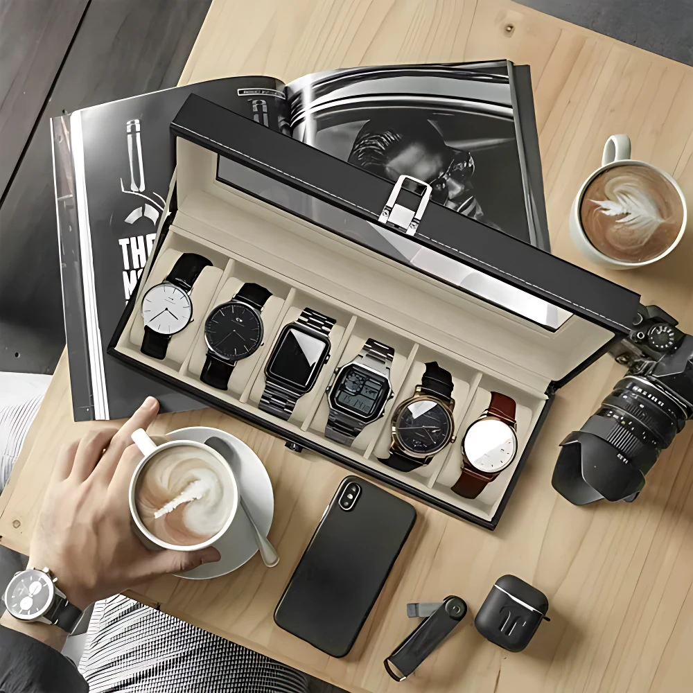 Relojero X6 negro con tapa de vidrio, abierto sobre mesa de madera, mostrando seis compartimentos con relojes de diferentes estilos y colores. Incluye teléfono móvil, cámara, taza de café y revista al fondo.