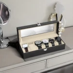 Relojero negro con tapa de vidrio, abierto y mostrando seis relojes de pulsera en compartimentos individuales sobre superficie clara.