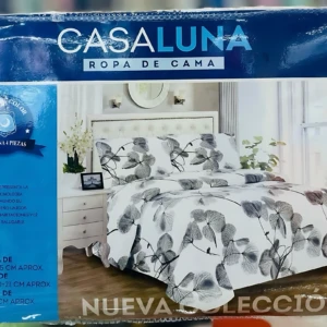 Juego de sábanas Casaluna de 4 piezas con estampado de hojas grises sobre fondo blanco. Se ve montado en cama blanca junto a mesita de noche. 203 x 235 cm.