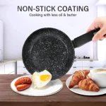 Sartén antiadherente negra con textura moteada, siendo volteada para servir un huevo frito sobre un plato con salchichas, en una mesa de madera.