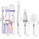 Set de tres utensilios de cocina en empaque: cuchillo de torta con hoja dentada, espátula de servir y rodillo para pizza, todos con mango acrílico transparente y detalles metálicos. Medidas visibles: 33.5 cm de altura total, 17 cm de ancho del empaque, 2.3 cm de grosor de hoja, 5 cm de ancho de espátula, 6.6 cm de diámetro del rodillo. Peso total: 162.5 g.