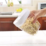 Mano sosteniendo recipiente transparente con tapa blanca, lleno de granos secos, sobre encimera de cocina con lavamanos y frutas al fondo.