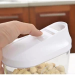 Mano cerrando tapa blanca de recipiente transparente con pistachos, sobre encimera de cocina con gabinetes de madera al fondo.