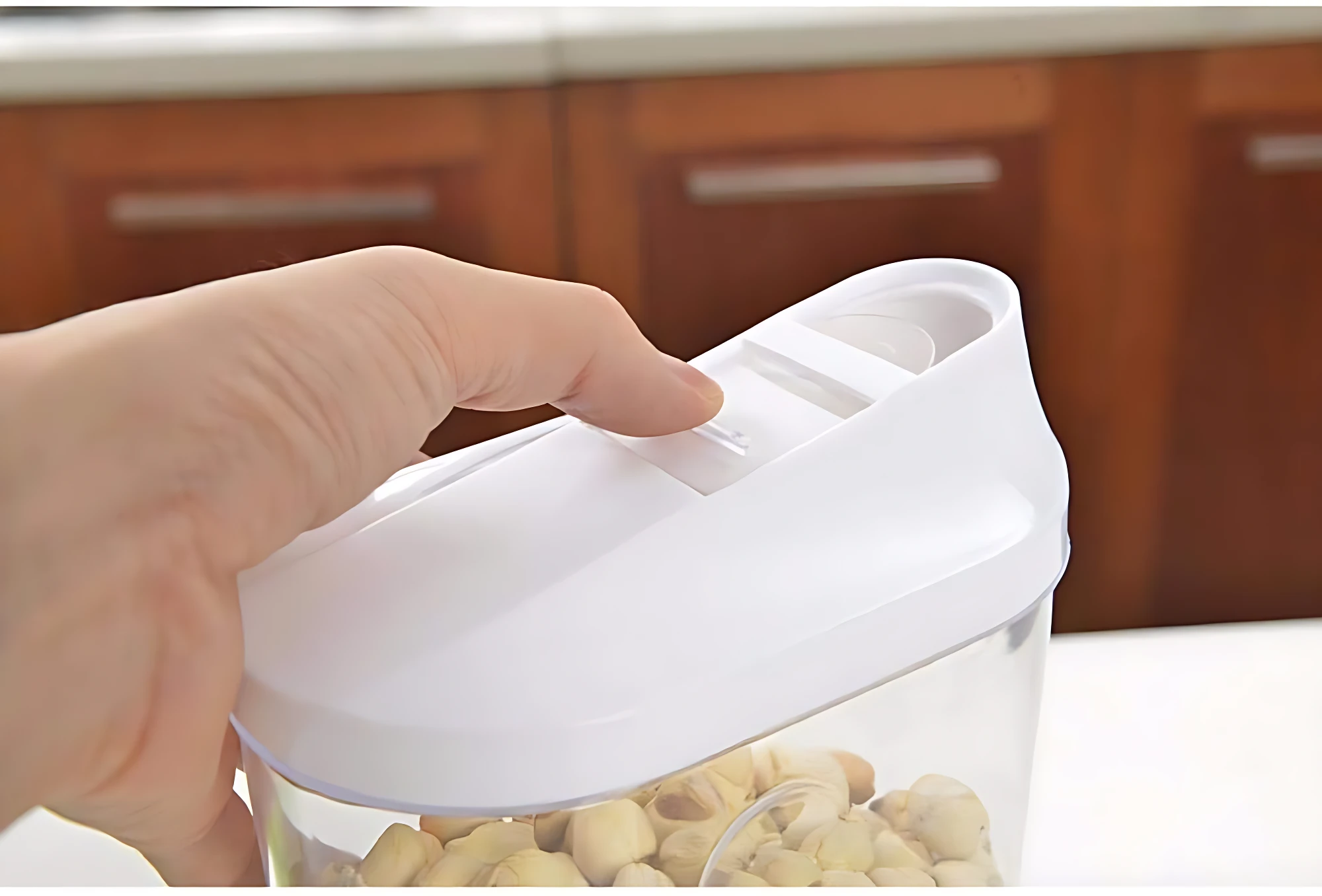 Mano cerrando tapa blanca de recipiente transparente con pistachos, sobre encimera de cocina con gabinetes de madera al fondo.