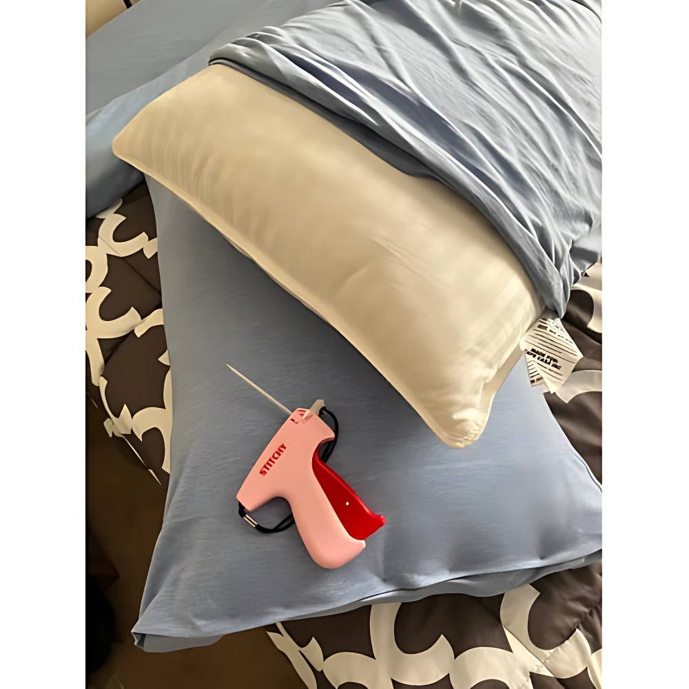 Pistola de puntadas Stitchy en color rosa con mango rojo, sobre funda de almohada azul claro y cojín beige, sobre cama con sábanas estampadas.