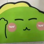 Tapete absorbente con diseño de animalito verde sonriente, mostrando expresión feliz con mejillas rosadas y mano levantada, ideal para decorar y proteger suelos en interiores.