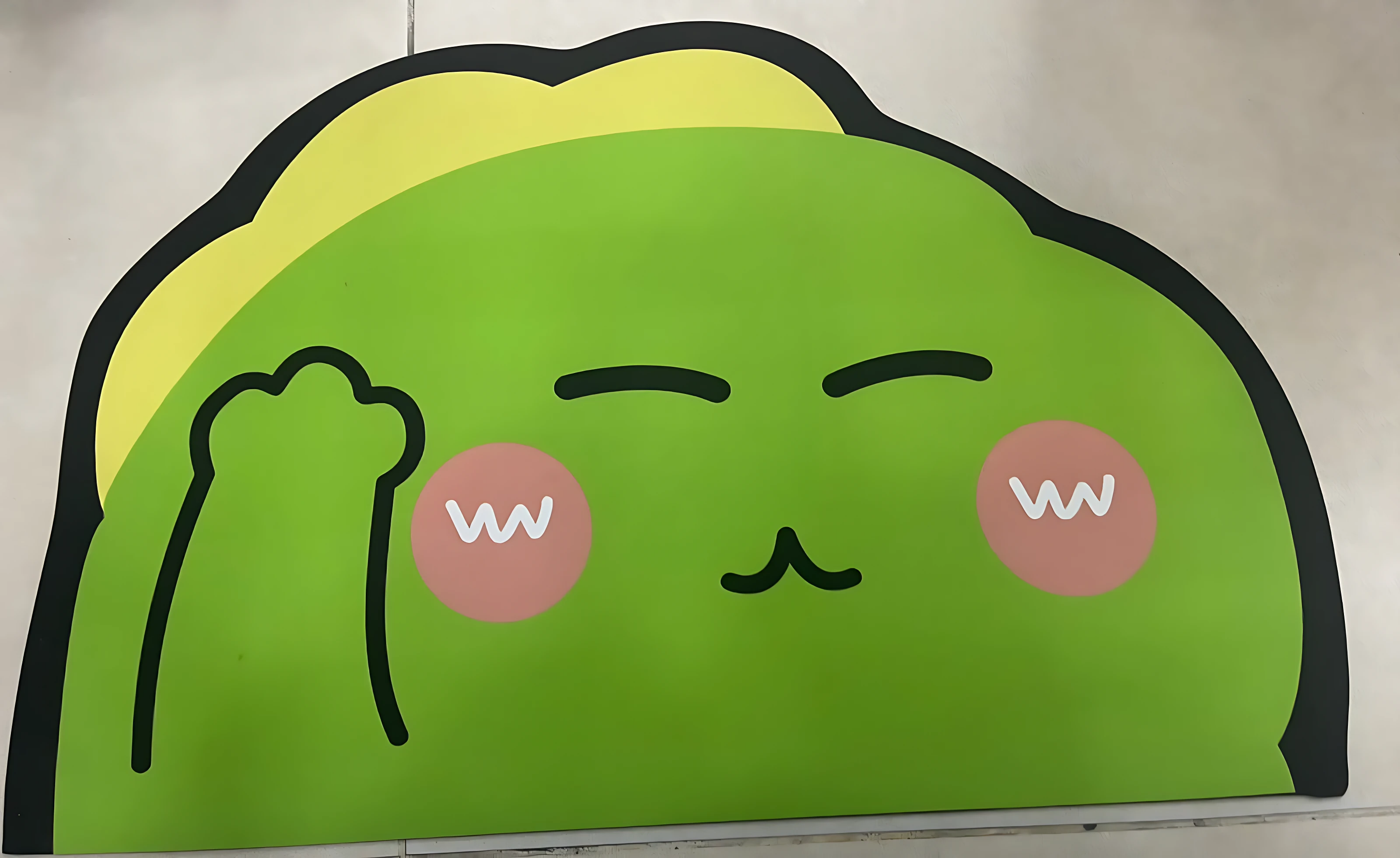 Tapete absorbente con diseño de animalito verde sonriente, mostrando expresión feliz con mejillas rosadas y mano levantada, ideal para decorar y proteger suelos en interiores.