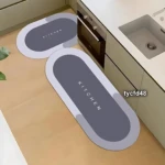 Dos tapetes absorbentes de cocina con diseño ovalado en gris y borde claro, uno pequeño y otro largo, ambos con la palabra "KITCHEN" impresa en el centro, colocados sobre un piso de madera clara junto a una encimera blanca con fregadero.