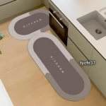 Dos tapetes absorbentes de cocina en tonos grises con la palabra Kitchen en relieve, dispuestos en el suelo junto a un fregadero y horno.