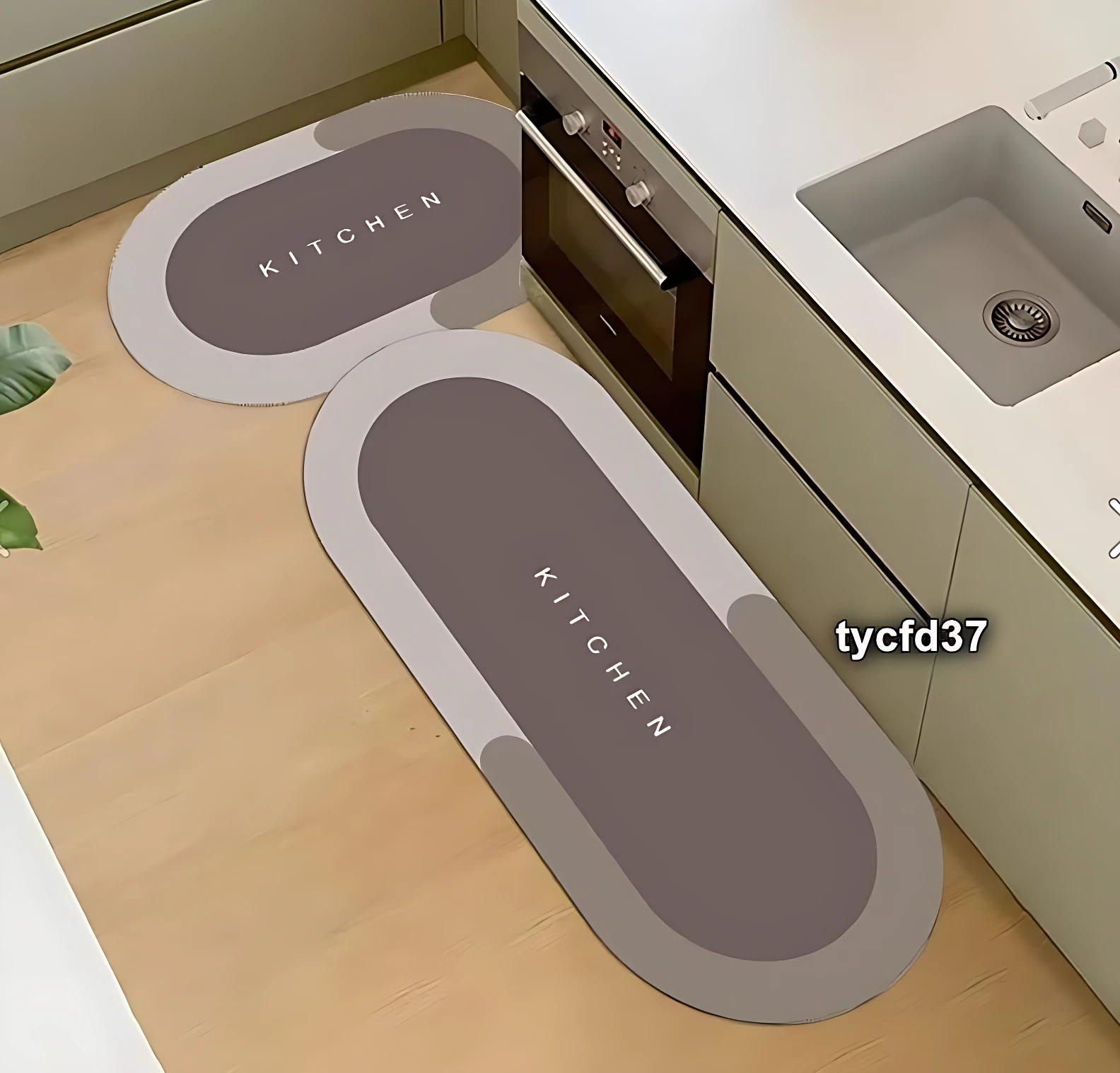 Dos tapetes absorbentes de cocina en tonos grises con la palabra Kitchen en relieve, dispuestos en el suelo junto a un fregadero y horno.