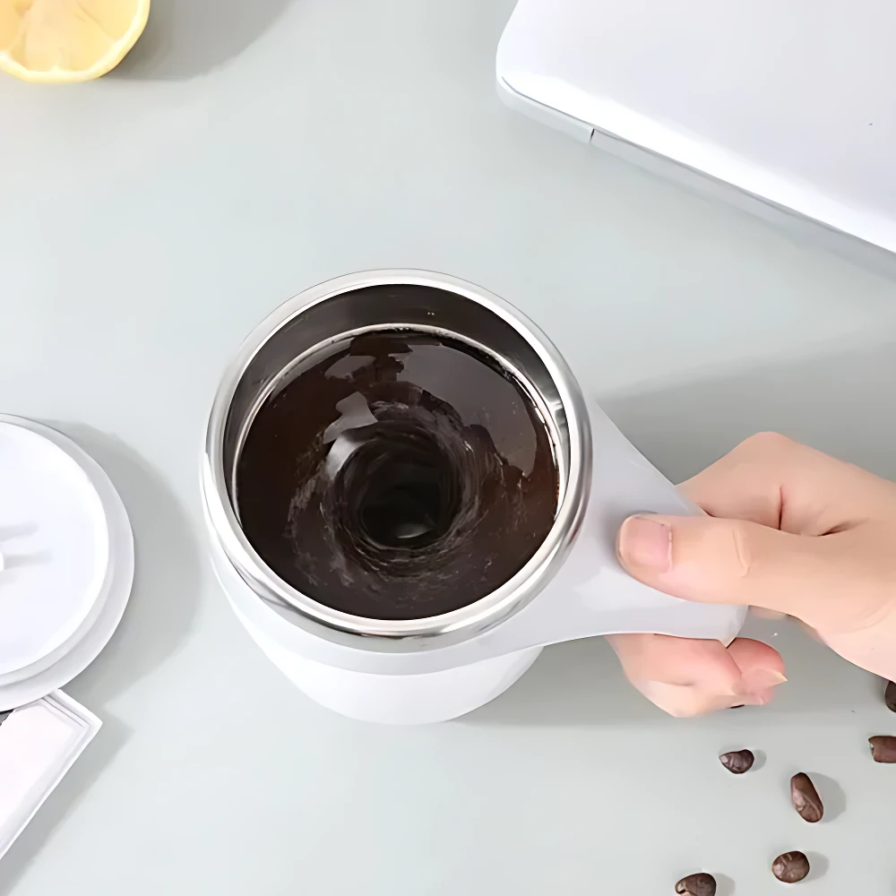 Taza mezcladora automática giratoria en color blanco con líquido oscuro, vista superior, mano sosteniendo la taza sobre superficie clara.