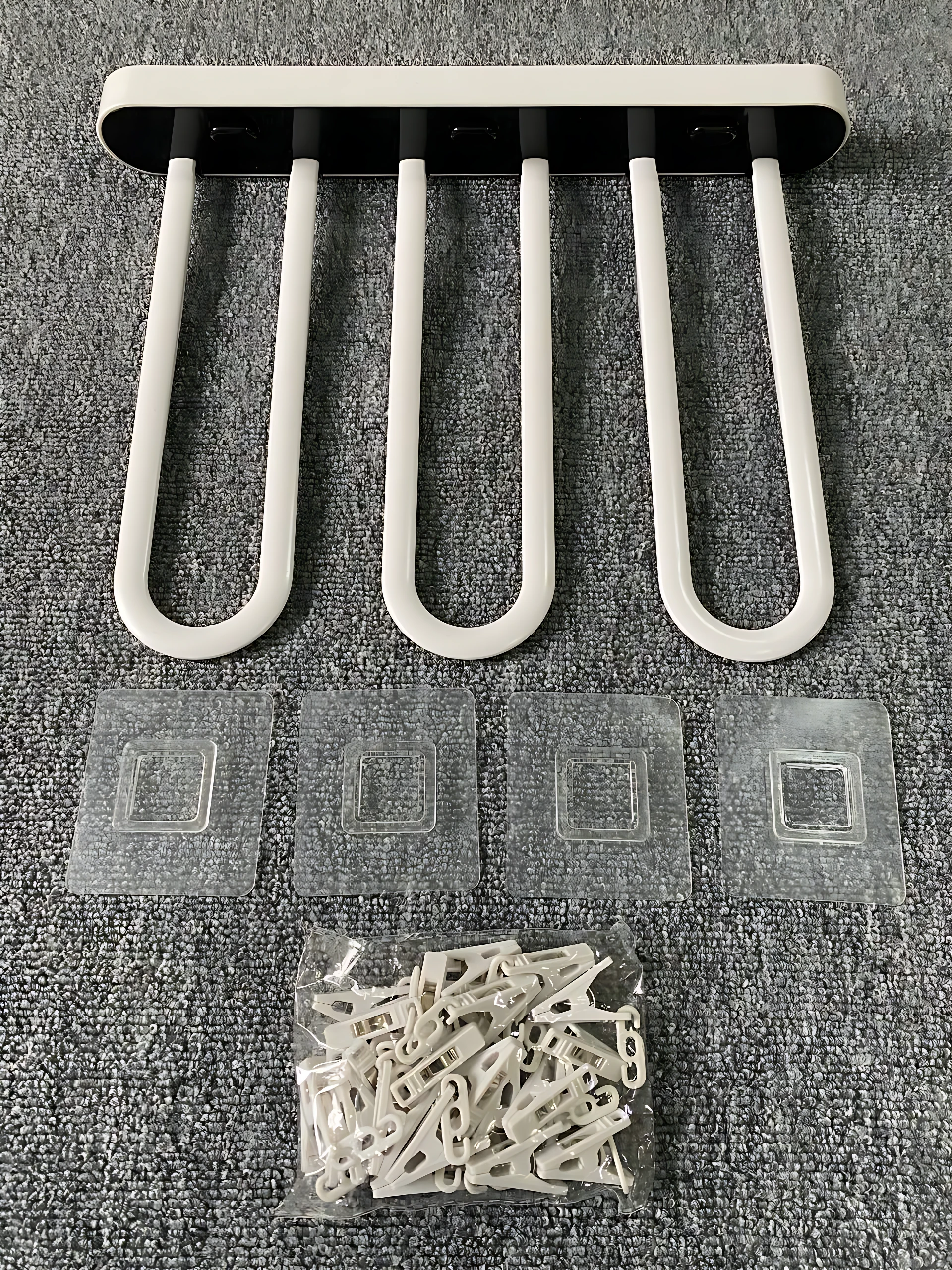 Tendedero plegable de plástico blanco con cinco ganchos en forma de U, acompañado de cuatro adhesivos y un paquete de clips de sujeción, sobre superficie texturizada gris.