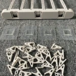 Tendedero plegable de plástico blanco con cuatro ganchos, cuatro adhesivos y múltiples pinzas de secado dispuestas sobre superficie gris texturizada.