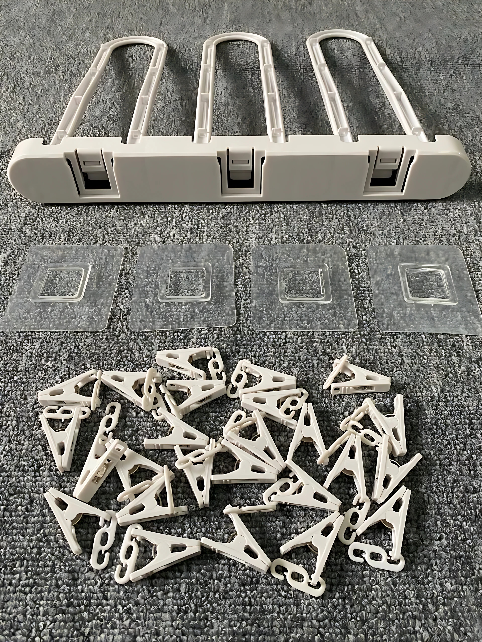Tendedero plegable de plástico blanco con cuatro ganchos, cuatro adhesivos y múltiples pinzas de secado dispuestas sobre superficie gris texturizada.