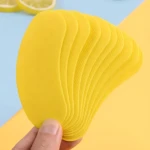 Mano sosteniendo tiras desodorantes amarillas para calzado, con textura ondulada y acabado mate, sobre fondo azul y amarillo con rodajas de limón al fondo.