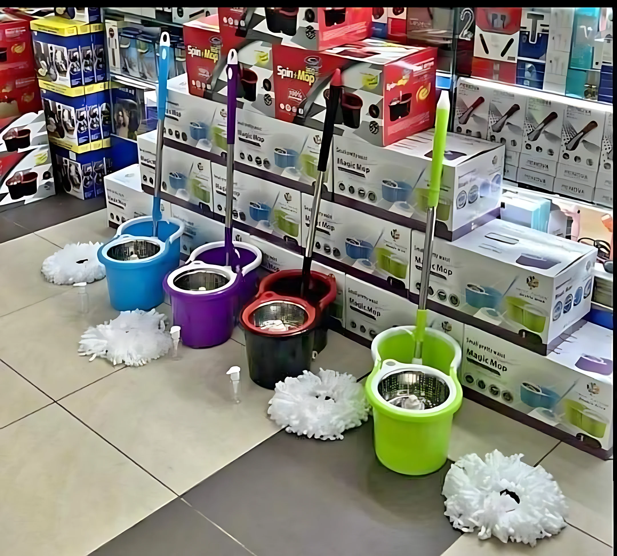 Varios traperos giratorios con tambor metálico en colores azul, morado, rojo, negro y verde, dispuestos en tienda con mopas blancas y cajas de repuesto al fondo.