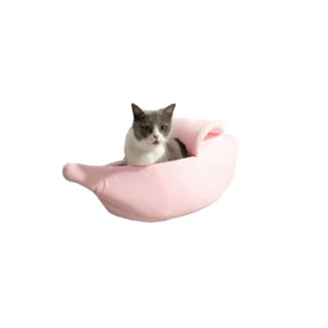 Cama en forma de banano color rosa con gato gris y blanco sentado dentro, sobre fondo blanco.
