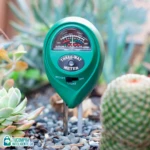 Medidor de suelo verde con tres puntas metálicas, para medir humedad, luz y pH. Se inserta en tierra entre suculentas y piedras decorativas.