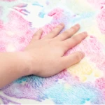 Mano sobre alfombra felpudo luminosa con patrones de luna y estrellas en tonos pastel rosa, azul y amarillo.