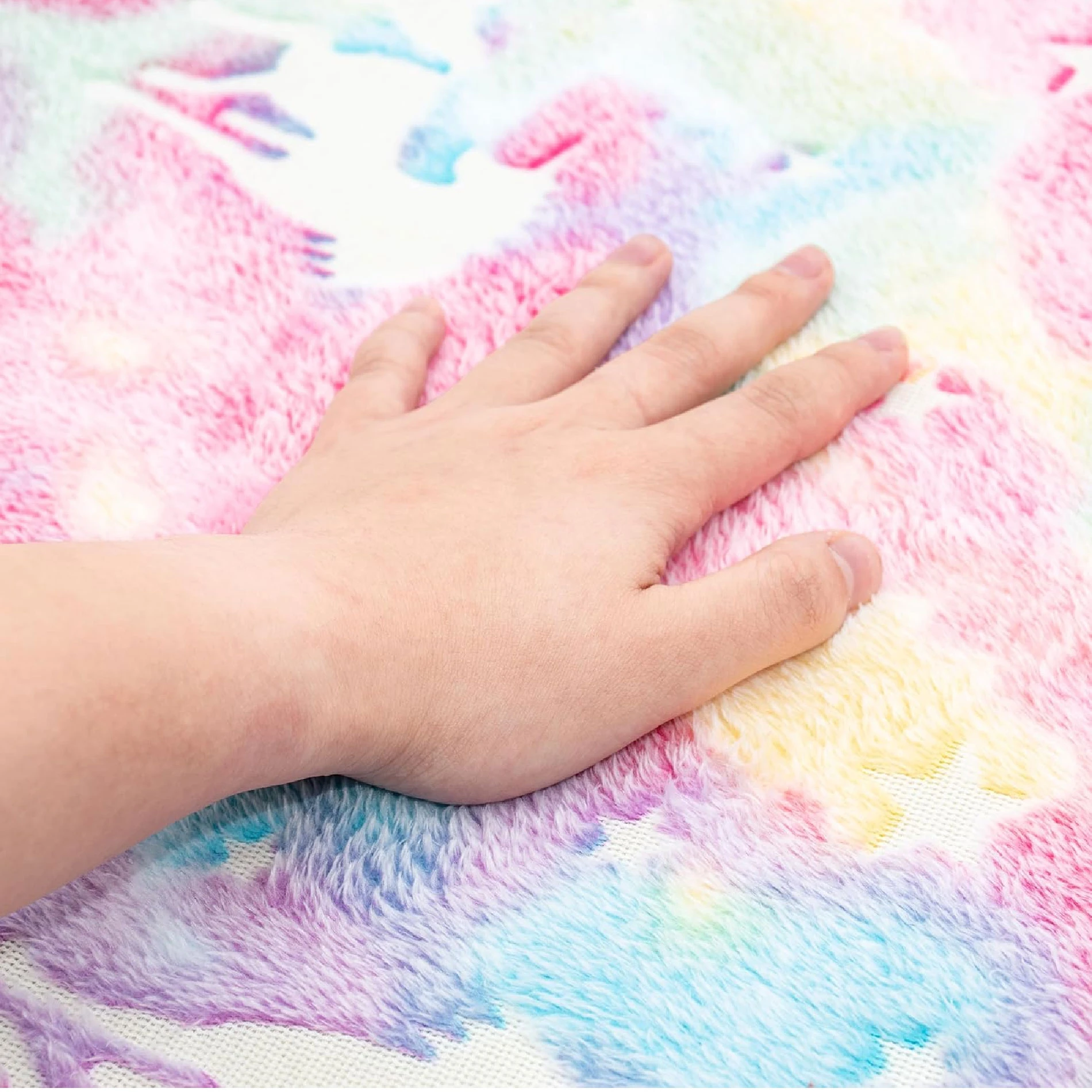 Mano sobre alfombra felpudo luminosa con patrones de luna y estrellas en tonos pastel rosa, azul y amarillo.
