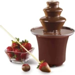 Fuente de chocolate eléctrica mini con cascada continua, color marrón oscuro, en uso con fresas y palillos de madera sobre fondo blanco.