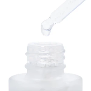 Dropper de vidrio con gota de suero transparente sobre boca de frasco blanco, mostrando textura líquida y aplicación directa para rostro.