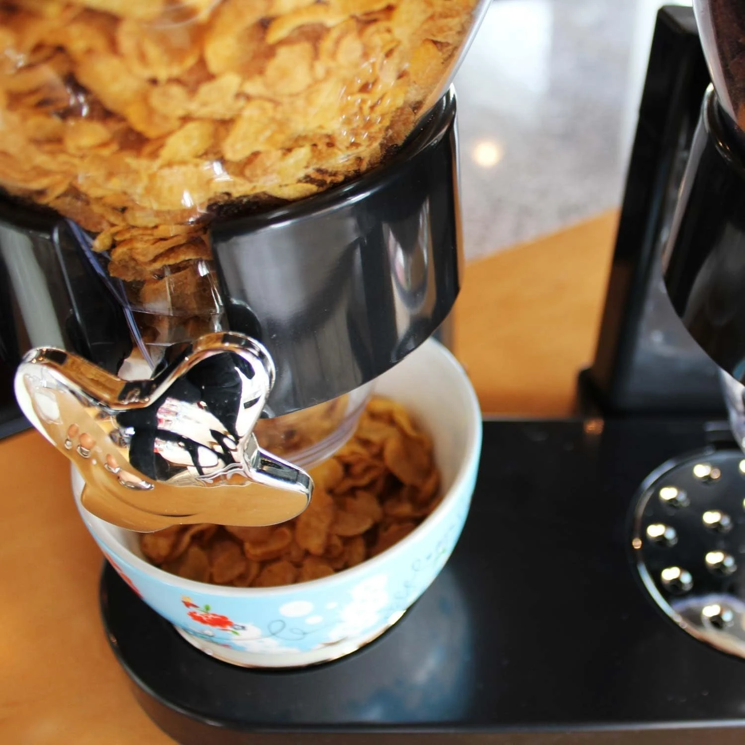 Dispensador de cereales de 2 litros con tapa transparente y dispensador manual en metal cromado, vertiendo granos dorados en taza azul con estampado floral sobre base negra.