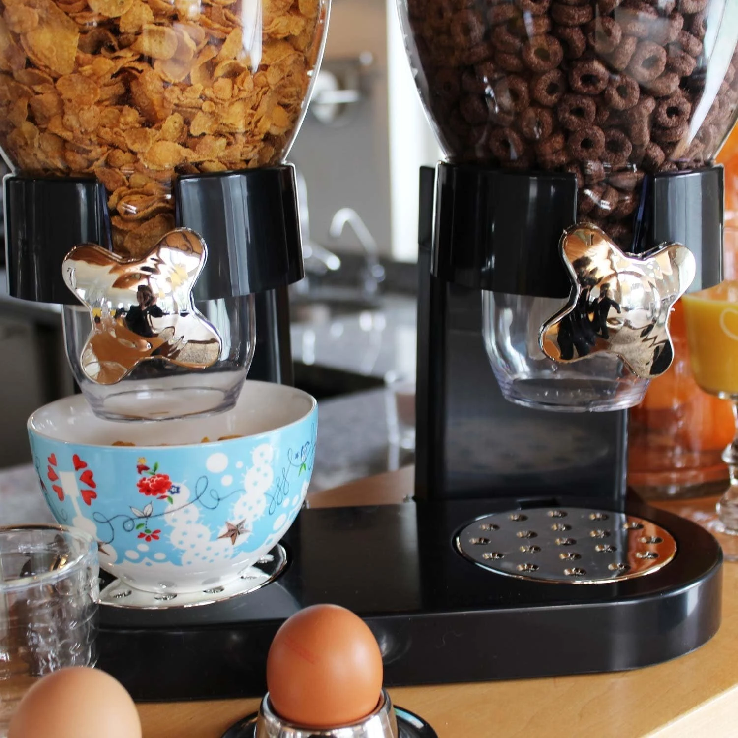 Dispensador de cereales de dos compartimentos con tapas transparentes, llenos de maíz y hojuelas de chocolate, sobre base negra con dispensador manual en forma de estrella. Incluye tazón floral azul debajo del grifo izquierdo.