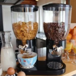 Dispensador de cereales de dos litros con dosificación manual, en negro, con dos recipientes transparentes llenos de maíz y chocolate, sobre encimera de cocina.