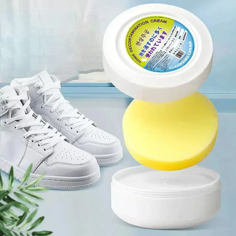 Contenedor de crema blanca con tapa amarilla y etiqueta multicolor, junto a un par de tenis blancos de alta costura sobre fondo claro con planta decorativa.