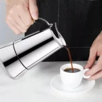 Cafetera espresso manual de acero inoxidable vierte café oscuro en taza blanca sobre plato, vista lateral con manos operando.