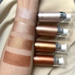 Cuatro iluminadores corporales Shimer en tonos dorados y bronce con acabado brillante, aplicados en el antebrazo junto a sus envases cilíndricos de cristal y metal.