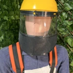 Casco amarillo con visera transparente y malla protectora negra, usado sobre camisa azul y delantal blanco, con correas naranjas visibles.