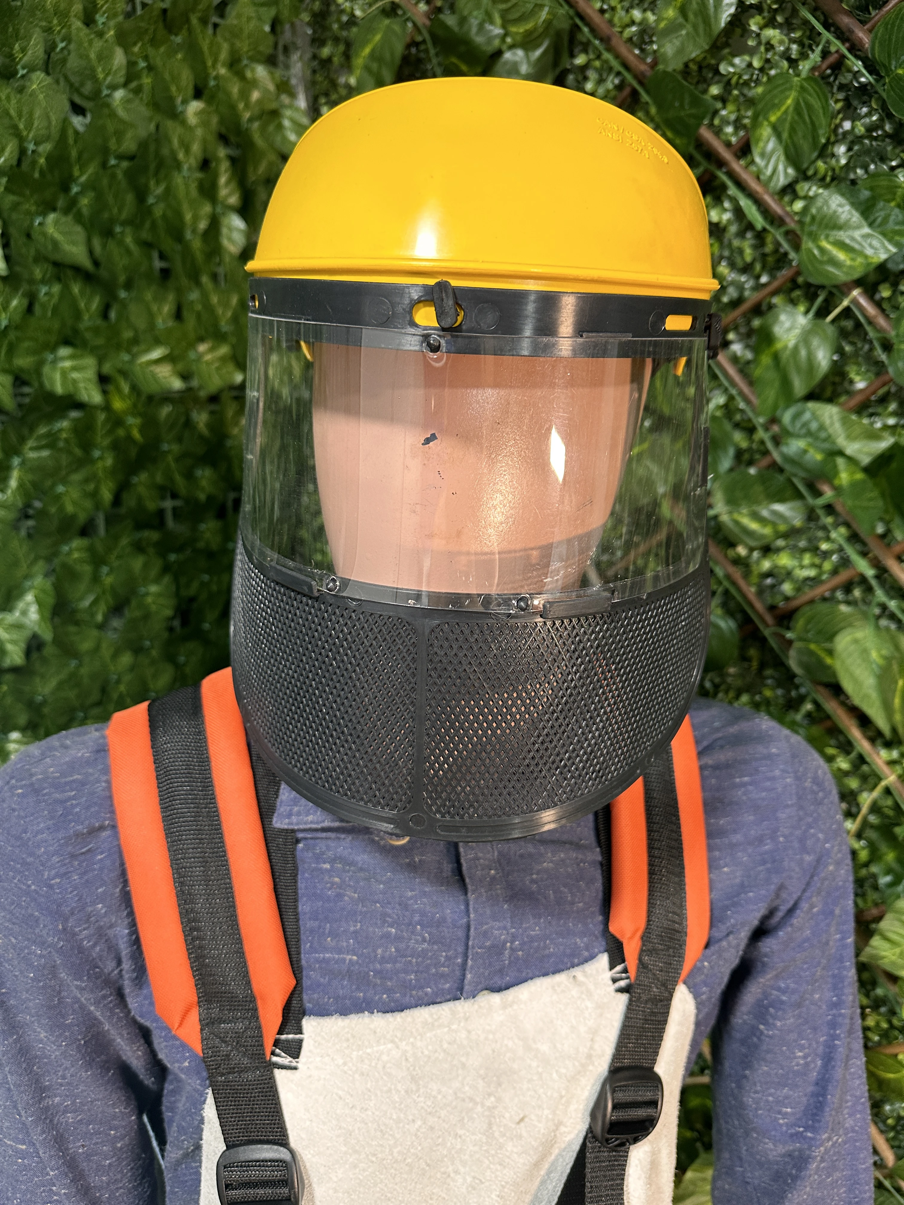 Casco amarillo con visera transparente y malla protectora negra, usado sobre camisa azul y delantal blanco, con correas naranjas visibles.