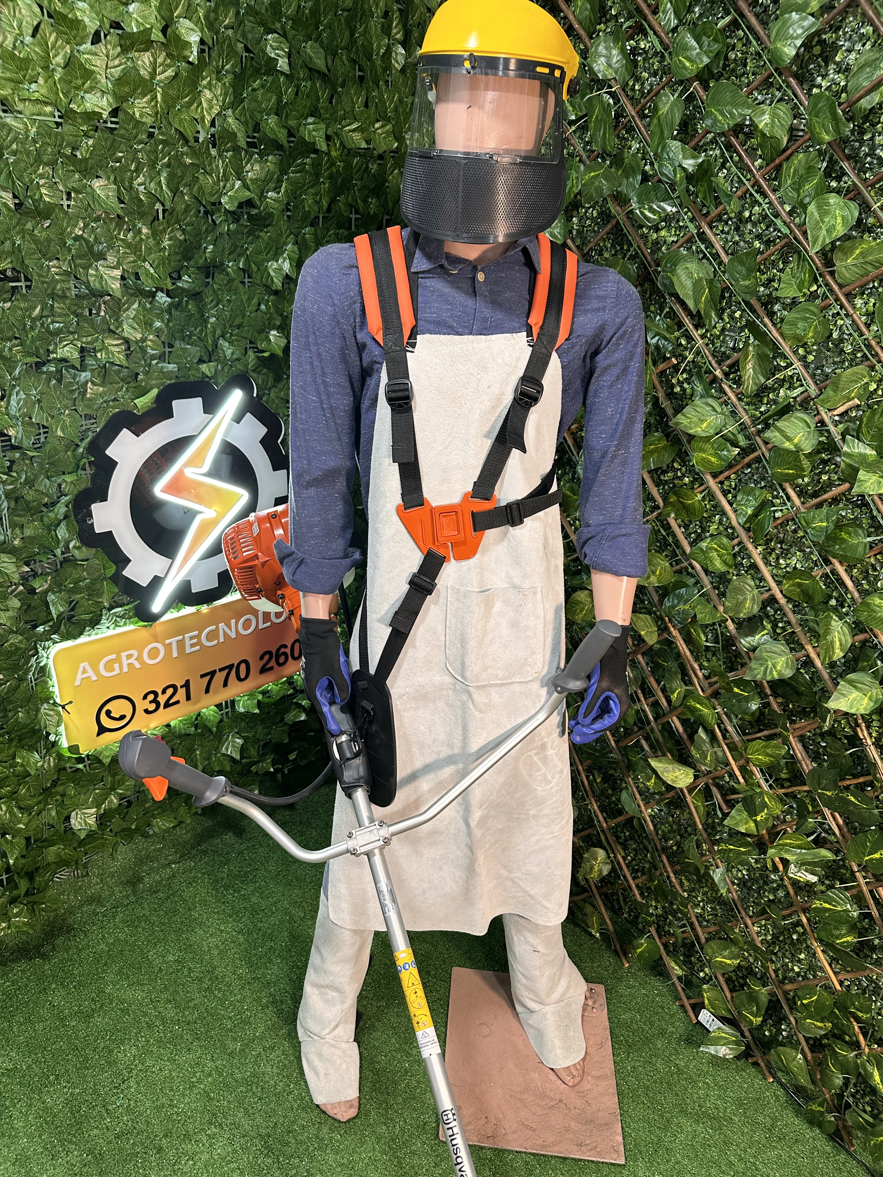 Maniquí con kit de protección integral para carnaza, incluyendo delantal beige, arnés naranja, casco amarillo con visera y guantes azules, sobre fondo de vegetación artificial.