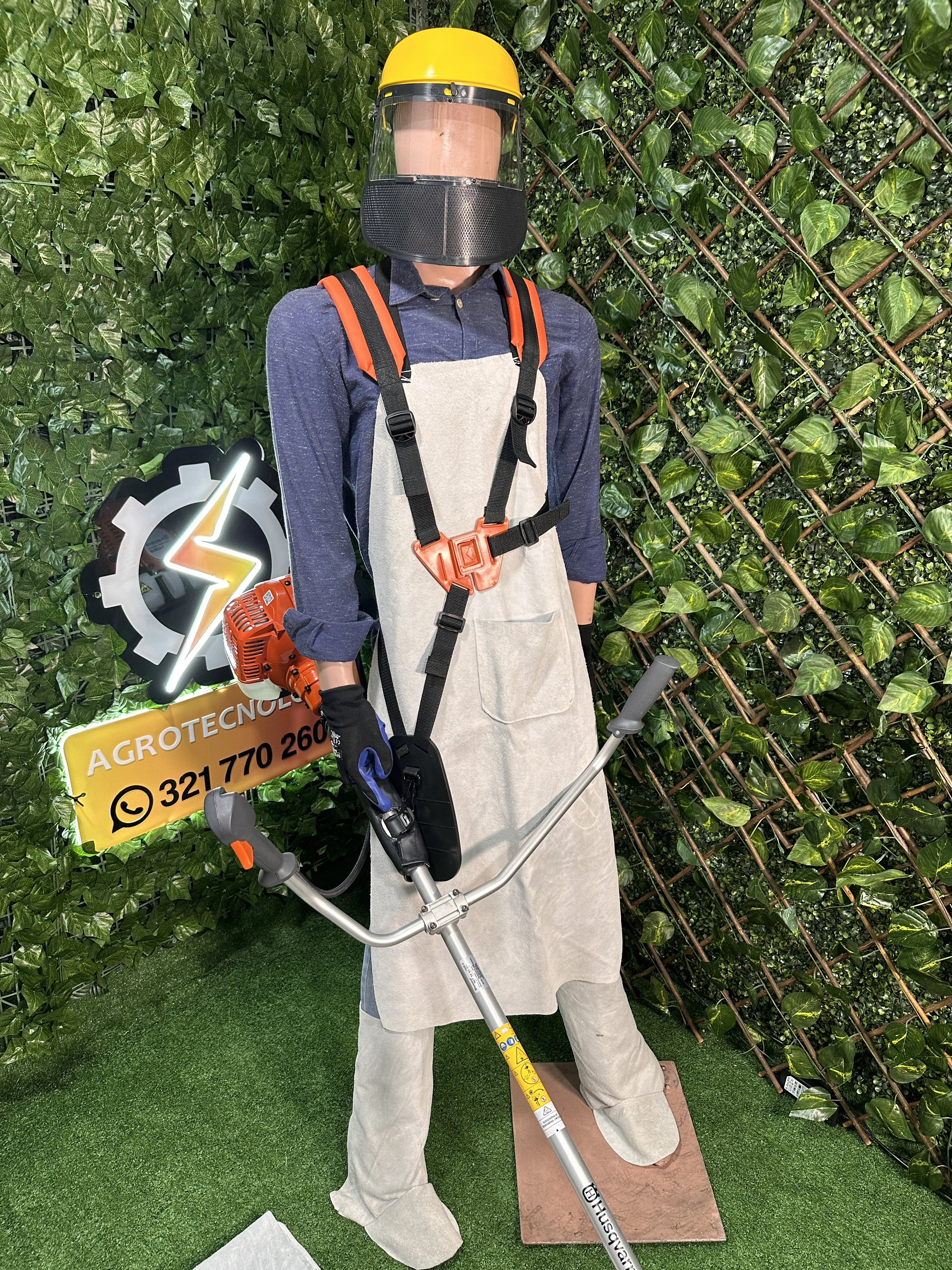 Maniquí con kit de protección integral para carnaza, incluyendo delantal beige, arnés naranja, casco amarillo con visera y guantes negros, sobre fondo verde artificial con logo Agrotecnia.