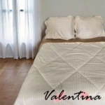 Edredón acanalado color crema con ojero, sobre cama doble con sábanas a juego, en habitación con cortinas blancas y piso de madera.