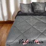 Edredón acanalado gris oscuro con ojero, textura de terciopelo y costuras diagonales, sobre cama doble con cojines a juego.