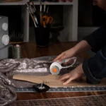 Cortador giratorio con hoja de titanio en uso sobre tela brillante y patrón de corte, sobre mesa de trabajo con accesorios de manualidades visibles.