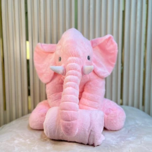 Peluche elefante rosa con cobija integrada, sentado sobre superficie blanca, textura suave y detalles en orejas y trompa visibles.