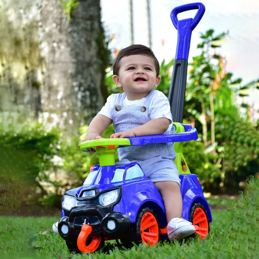Niño sonriente sentado en carro paseador azul con ruedas naranjas, mango ergonómico y barandilla verde, sobre césped al aire libre.