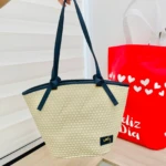 Bolso de mano Yute Ref Julieta en tejido natural color crema con asas azul marino, mostrando etiqueta frontal y fondo con bolsa roja decorada.