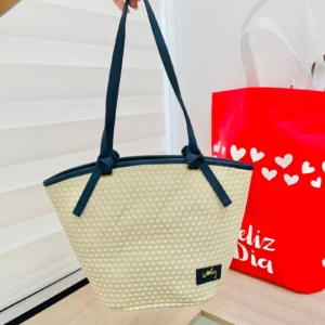 Bolso de mano Yute Ref Julieta en tejido natural color crema con asas azul marino, mostrando etiqueta frontal y fondo con bolsa roja decorada.