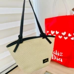 Bolso de mano tejido en yute natural con asas negras y detalles metálicos, ideal para llevar compras frescas.
