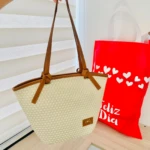 Bolso de mano tejido en yute natural con asas de cuero marrón, frente lisa y etiqueta visible. Se muestra junto a una bolsa roja con corazones blancos y el texto "Feliz Día".